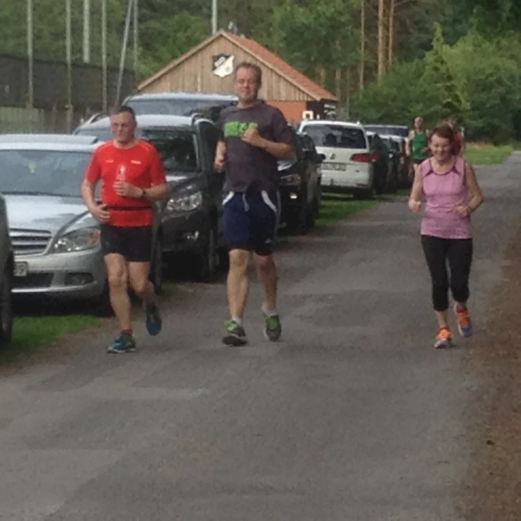 Drei Personen joggen auf einer Straße neben geparkten Autos im Grünen, Fitness und Outdoor-Aktivität, Sommerlauf.