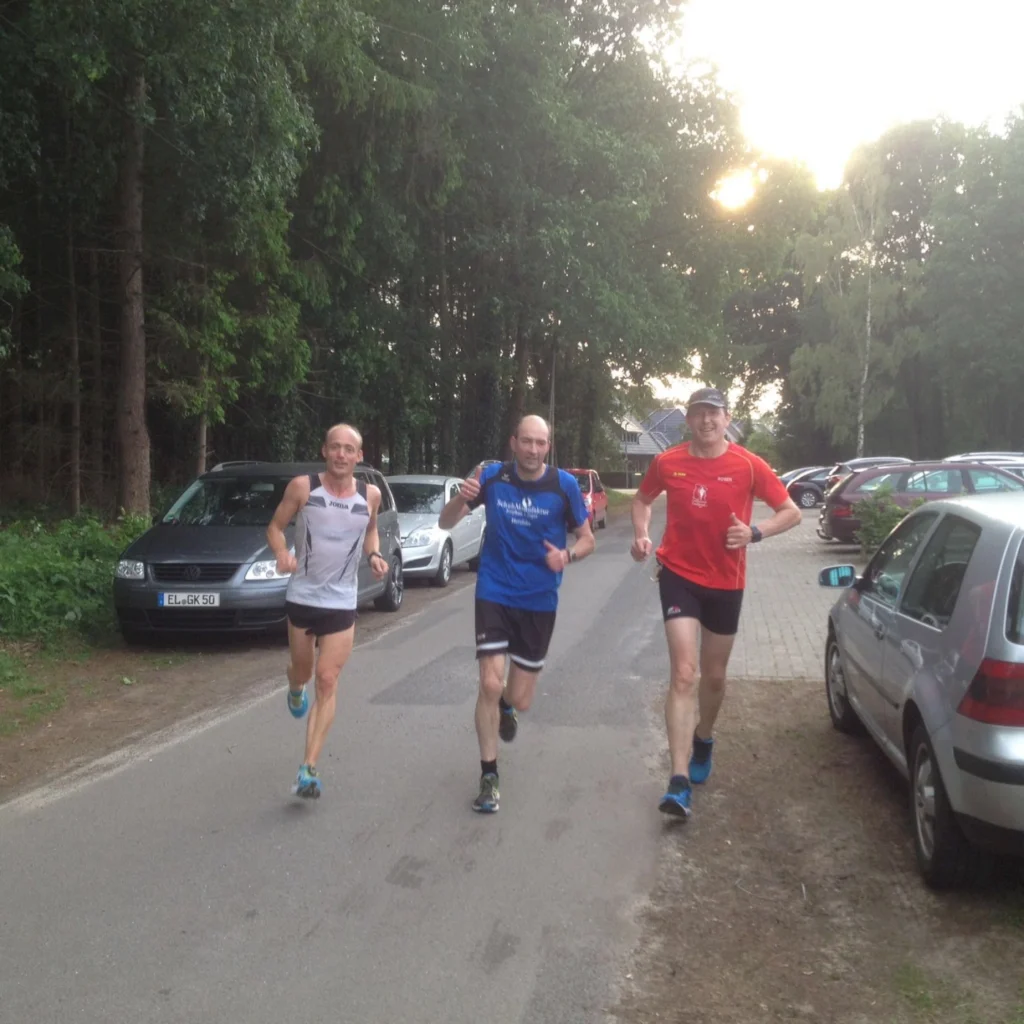 Drei Läufer joggen auf einem Weg, umgeben von Bäumen und parkenden Autos. Der erste Läufer trägt ein weißes Shirt, der zweite ein blaues, und der dritte ein rotes Shirt. Die Szene vermittelt eine sportliche Atmosphäre in der Natur.