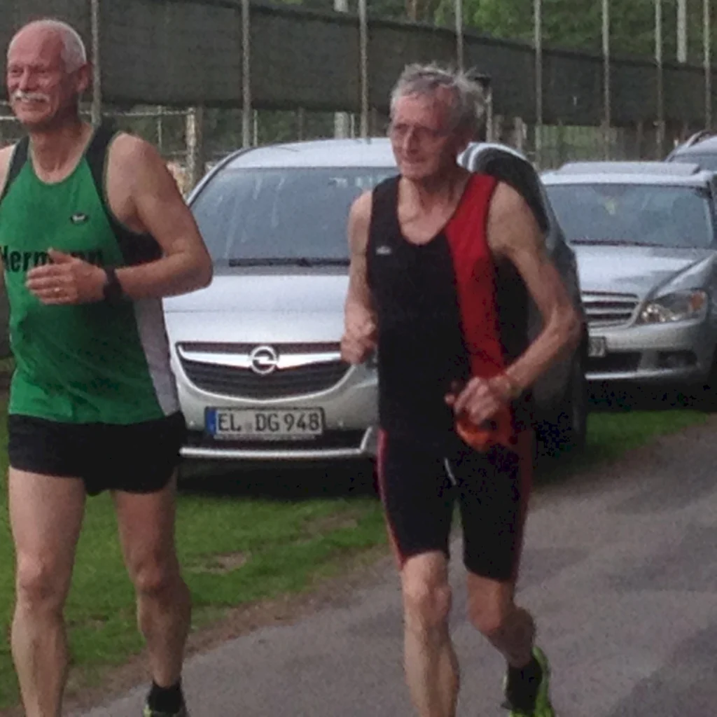 Zwei ältere Männer joggen auf einem Weg neben geparkten Autos, in sportlicher Laufkleidung und drücken Freude aus.