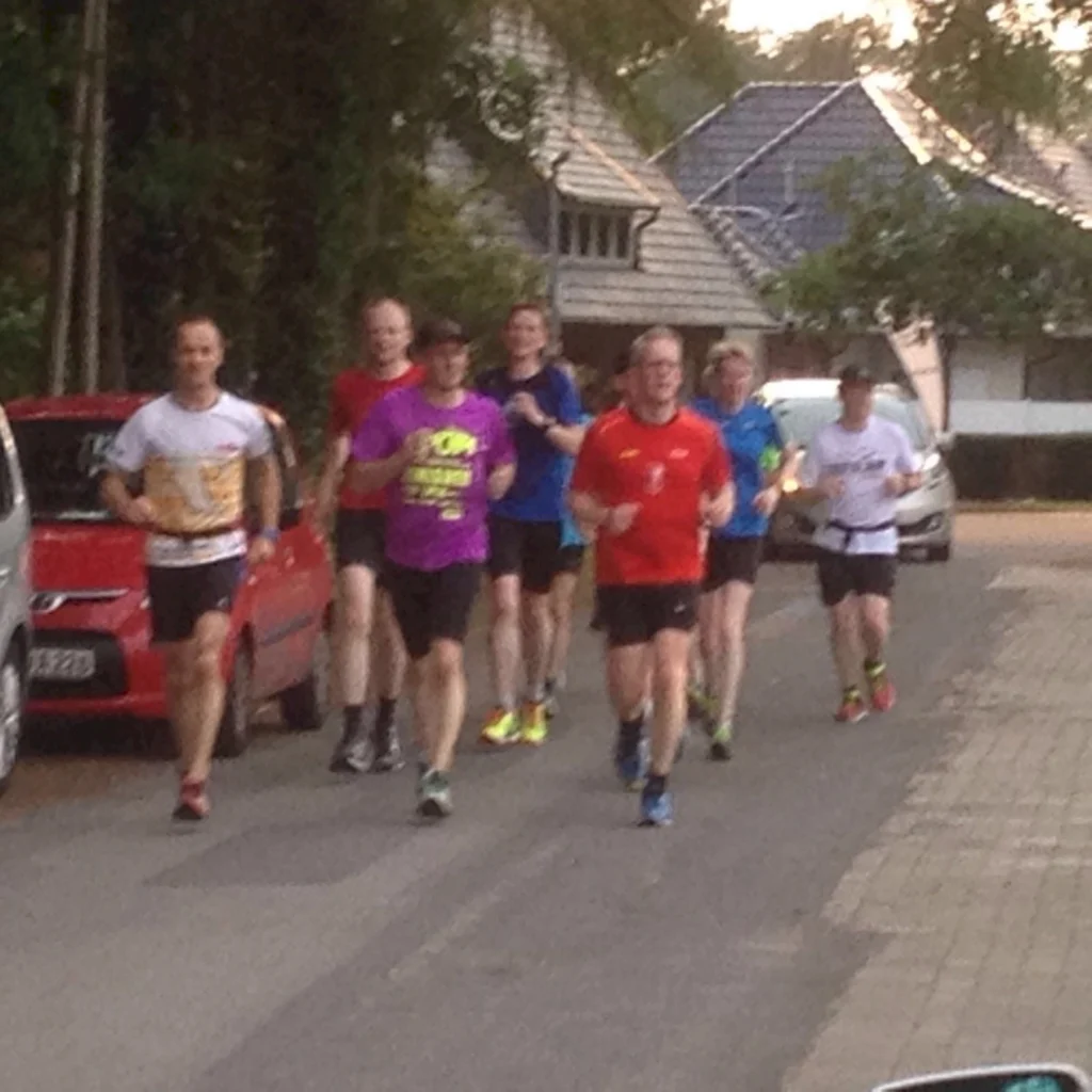 Gruppe von Läufern joggt auf einer Vorstadtstraße, entspannte Atmosphäre, bunte Sportkleidung, Bäume und Autos im Hintergrund.