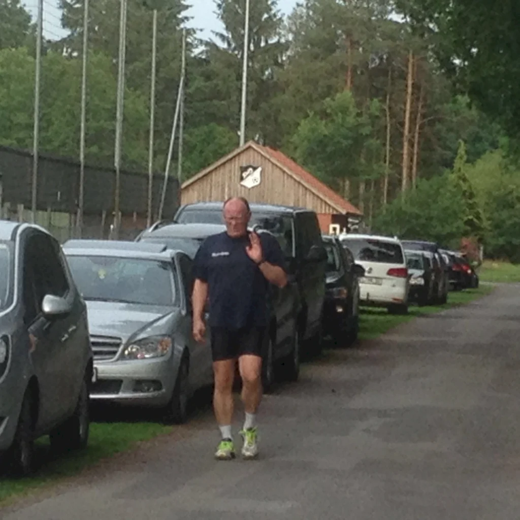 Ein Mann in Sportkleidung winkt beim Joggen auf einer Straße, gesäumt von parkenden Autos, mit einem Holzgebäude im Hintergrund.