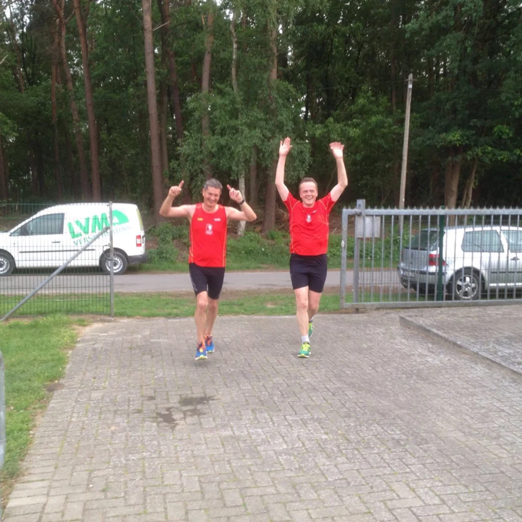 Zwei Männer in roten Sportoutfits jubeln beim Laufen im Freien, viele Bäume im Hintergrund.