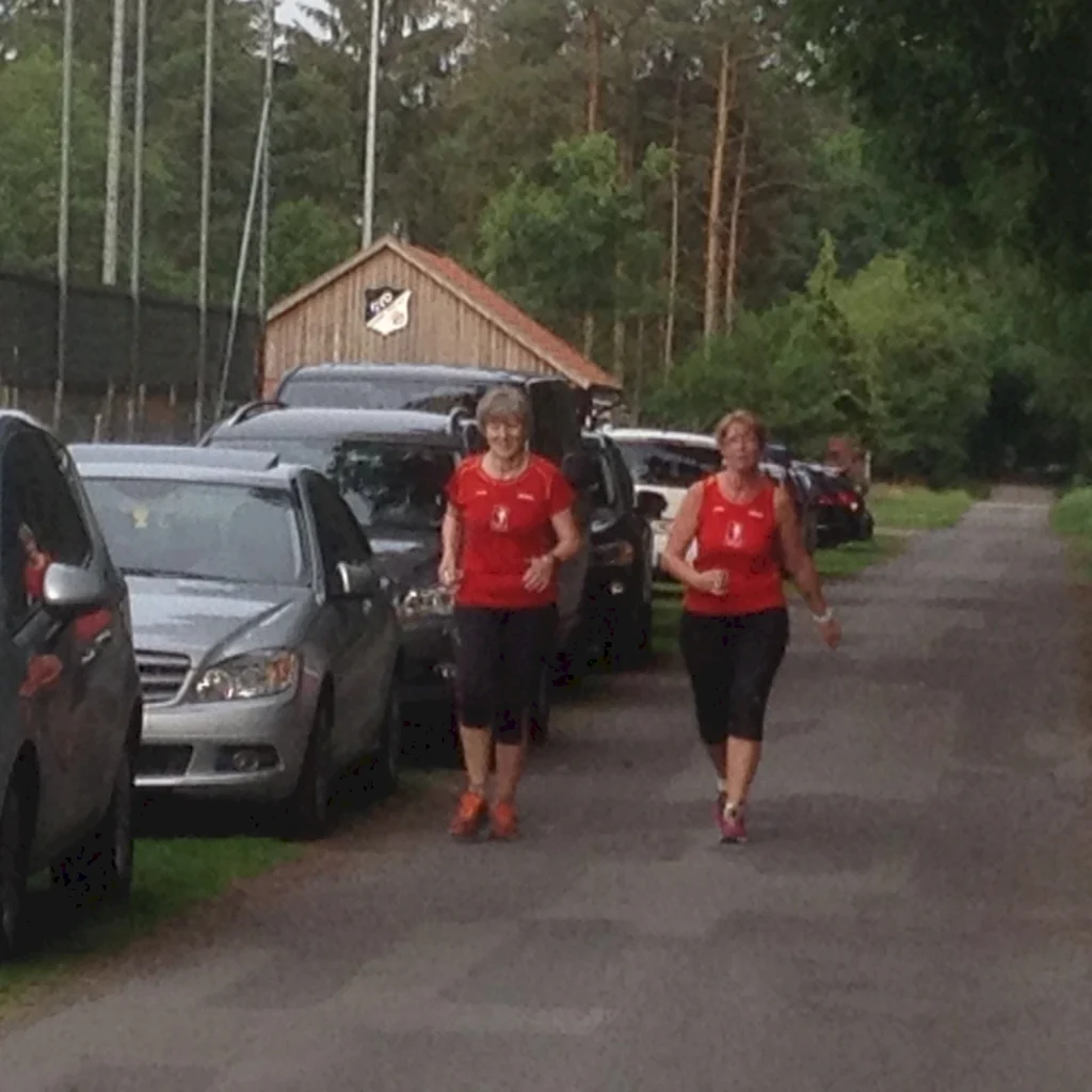 Zwei Frauen in roten Trikots joggen auf einem Weg entlang parkender Autos und grüner Bäume.