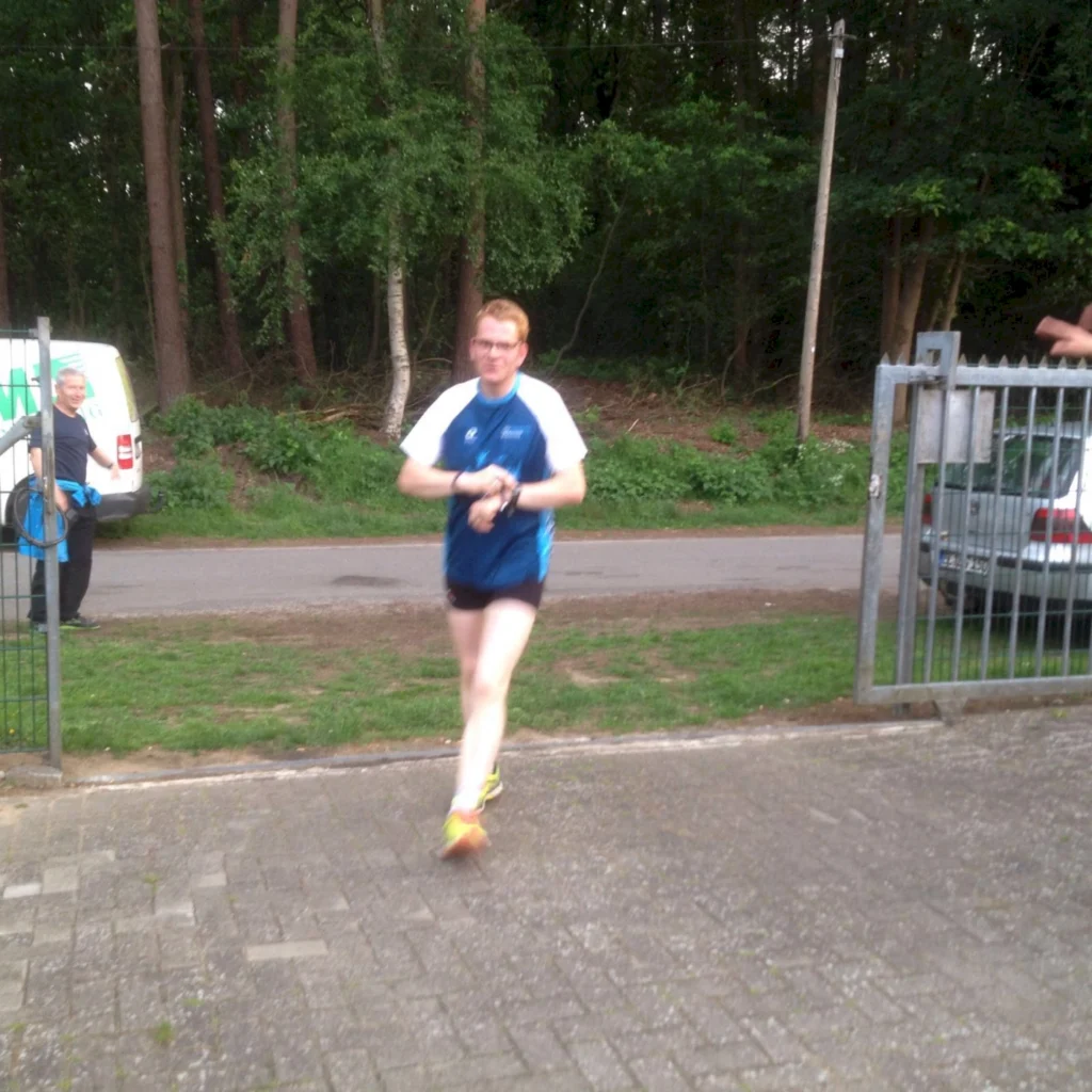 Läufer in Sportkleidung beim Joggen auf einem Weg, im Hintergrund ein Mann und ein Lieferwagen, umgeben von Bäumen und Grünflächen.