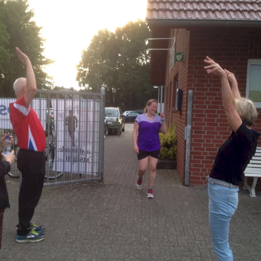 Frau in Sportkleidung beendet Getränkelauf, wird von zwei Menschen vor Sportgelände begrüßt.