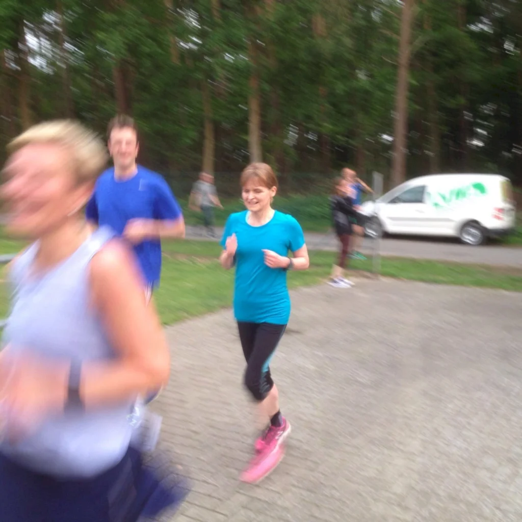 Läufer in einem Park bei einem Gruppenlauf, mit einer Frau in einem blauen T-Shirt und einer anderen Person im Hintergrund. Die Szene zeigt Bewegung und Fitnessaktivitäten im Freien.