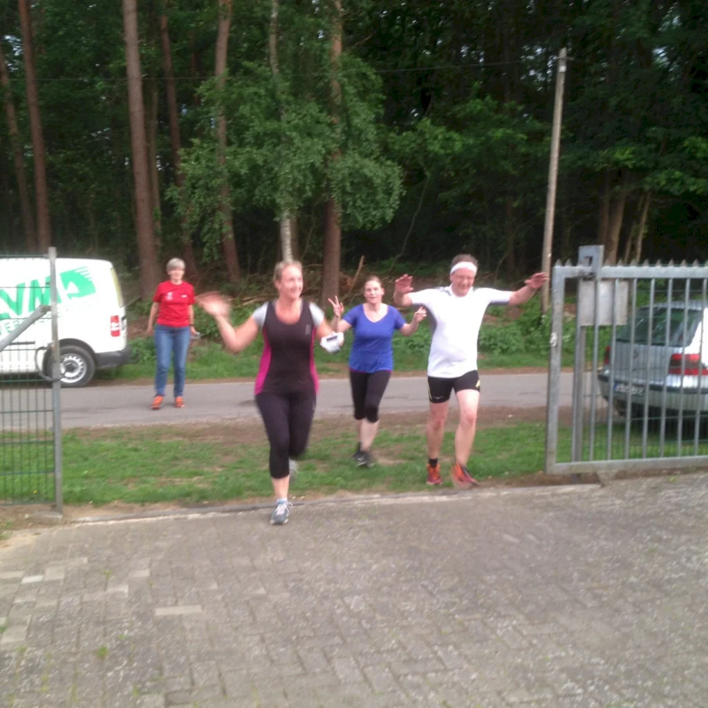 Gruppe von vier Personen beim Laufen im Freien, umgeben von Bäumen. Eine Frau in Sportkleidung führt die Gruppe an, während die anderen fröhlich folgen. Im Hintergrund ist ein Lieferwagen und ein geparktes Auto zu sehen, was auf eine sportliche Veranstaltung oder ein gemeinsames Training hinweist.