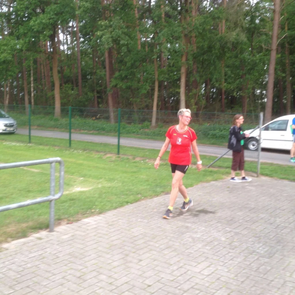 Frau in rotem Sportshirt spaziert auf einem Weg im Freien, Bäume und geparkte Autos im Hintergrund.