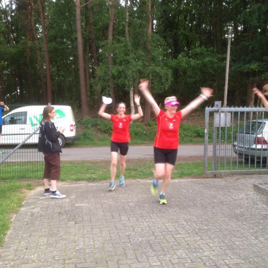 Zwei Frauen in roten Sporttrikots jubeln beim Laufen, während eine Zuschauerin am Rand applaudiert. Im Hintergrund sind Bäume und ein Fahrzeug sichtbar.