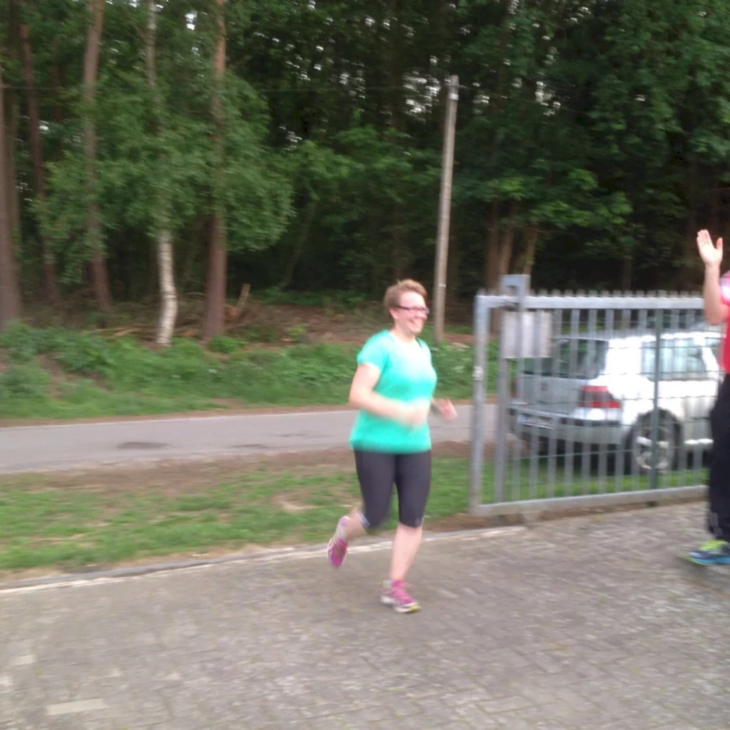 Frau joggt im Freien auf einem Weg neben einem Auto und einem Wald. Sie trägt Sportbekleidung und sieht zufrieden aus.