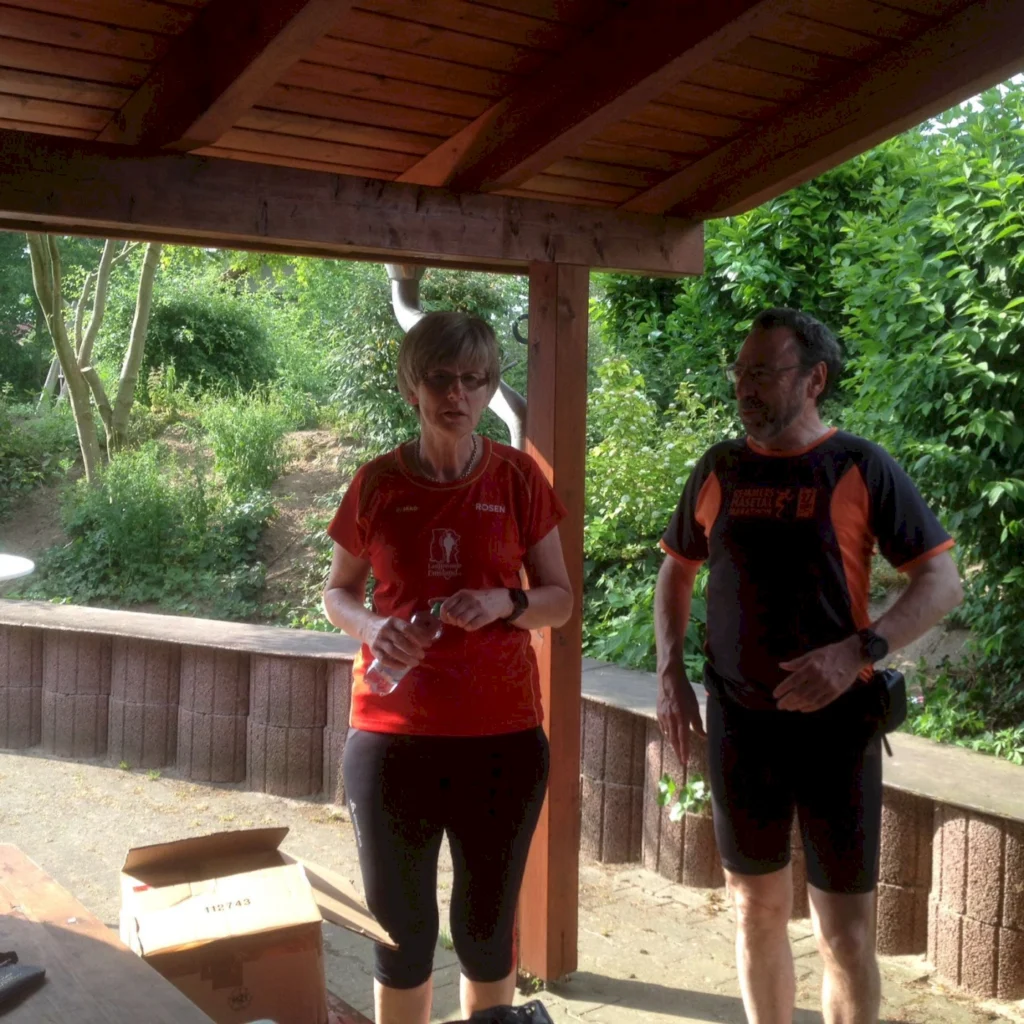 Zwei Personen in Sportkleidung unter einem Holzpavillon im Grünen, mit Bäumen und Pflanzen im Hintergrund.