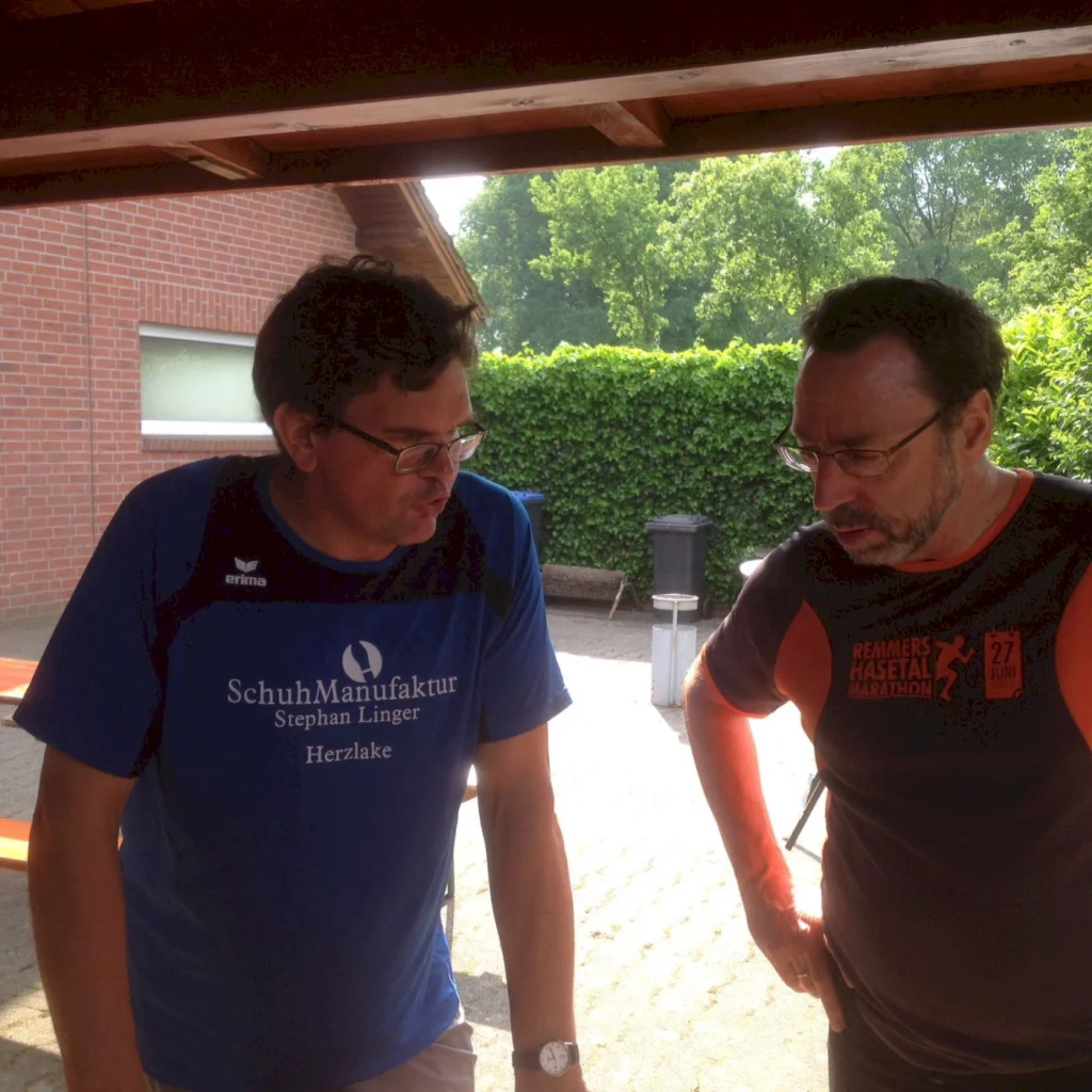 Zwei Männer im Gespräch im Freien, beide in T-Shirts, stehen unter einer Überdachung bei sonnigem Wetter.