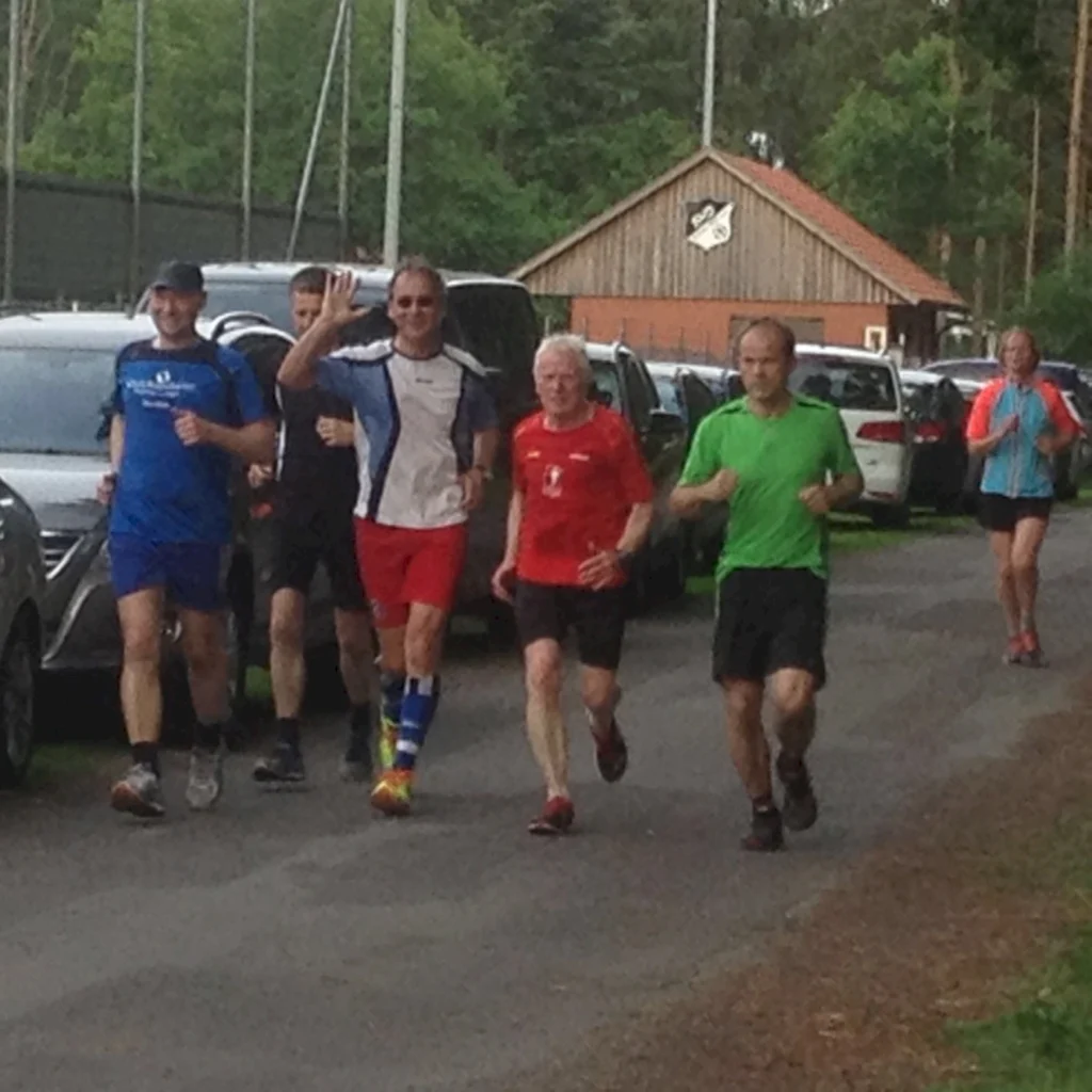 Gruppe von Läufern in sportlicher Kleidung joggt auf einem Weg entlang geparkter Autos, umgeben von Bäumen und einem Vereinsgebäude im Hintergrund.