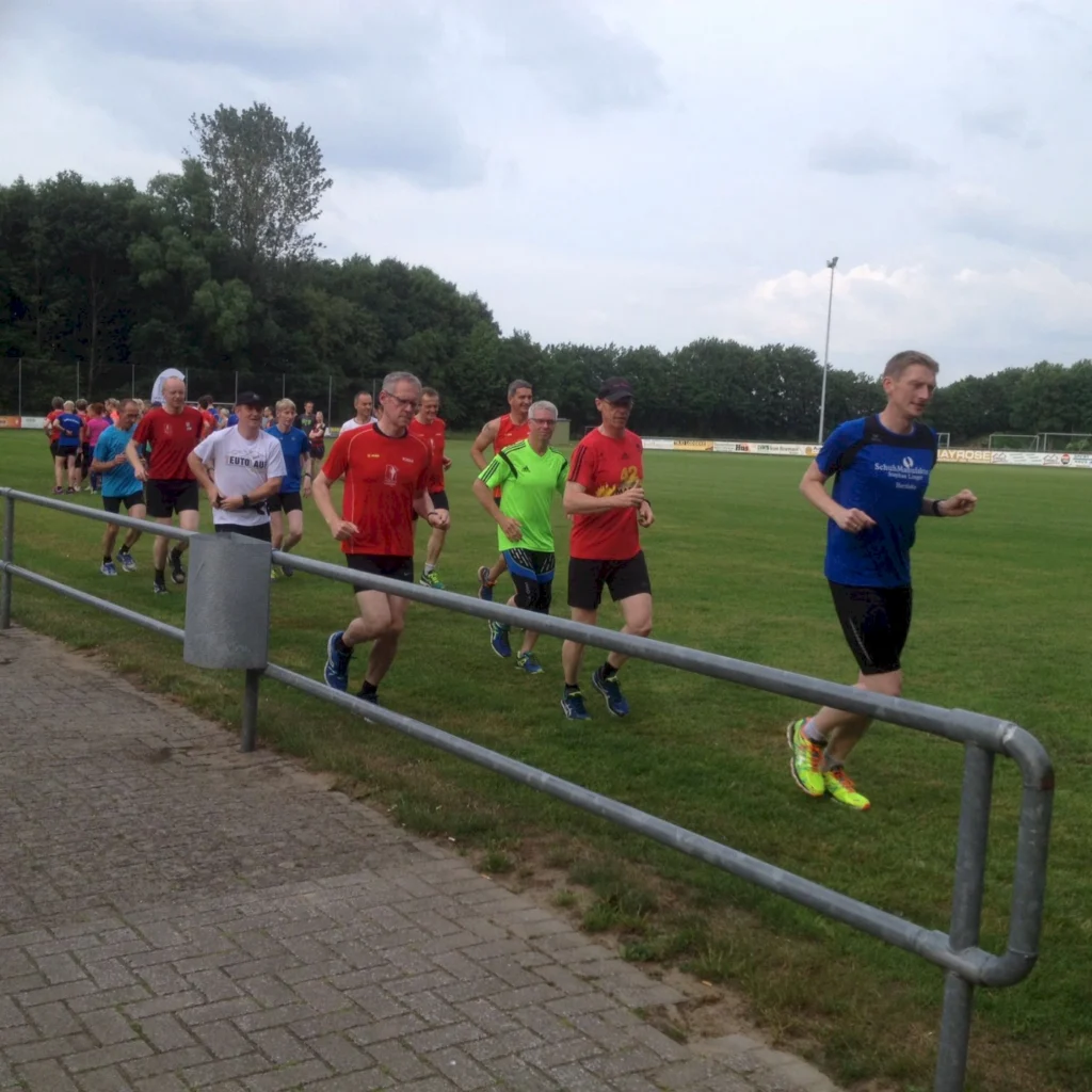 Gruppe von Läufern in bunter Sportkleidung trainiert auf grünem Rasen neben einer Laufbahn bei bewölktem Himmel.