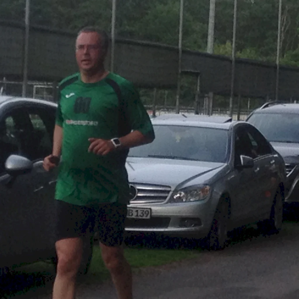 Mann joggt auf einem Gehweg neben geparkten Autos, trägt grünes Trikot und schwarze Shorts, Sportplatz im Hintergrund.