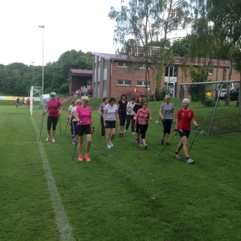 Gruppe von Menschen beim Nordic Walking auf einem Rasenplatz vor einem Gebäude im Freien.