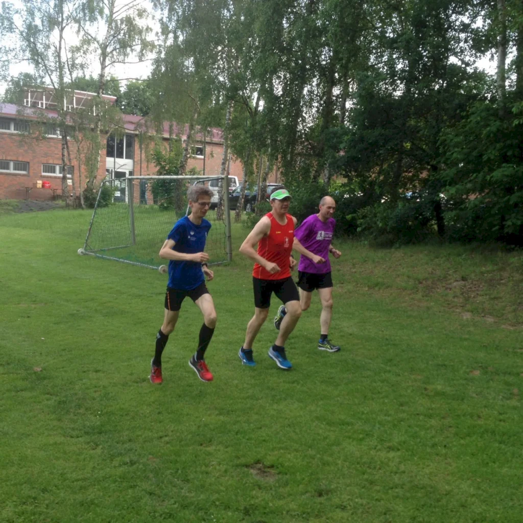 Drei Männer in Sportkleidung joggen auf einem grünen Rasenplatz. Im Hintergrund sind Bäume und ein Fußballtor sichtbar. Die Szene vermittelt eine aktive und gesunde Lebensweise.