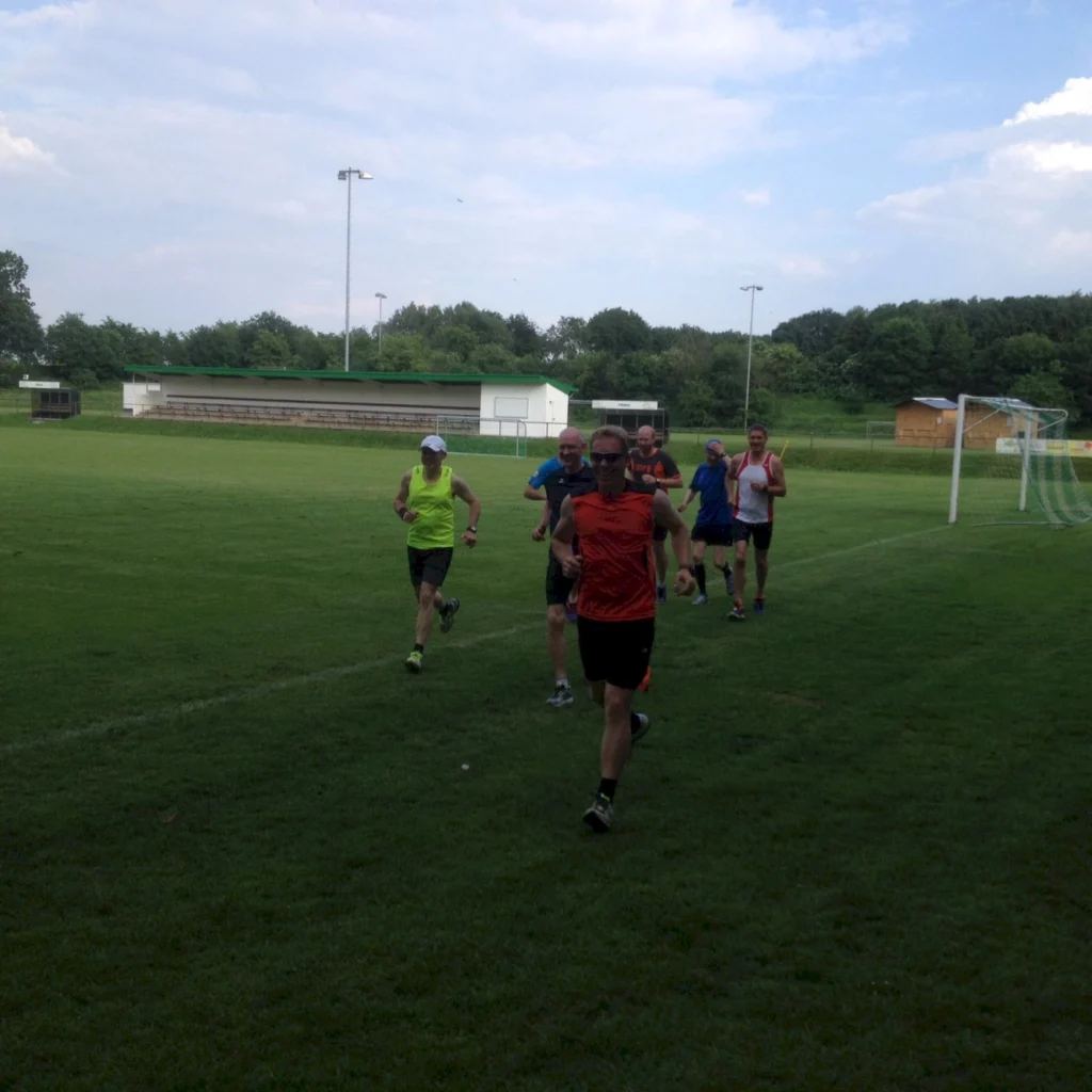 Gruppe von Männern beim Laufen auf einem Fußballplatz, umgeben von grünem Rasen und Bäumen im Hintergrund. Die Sportler tragen verschiedene Laufoutfits und zeigen eine aktive Trainingssituation.