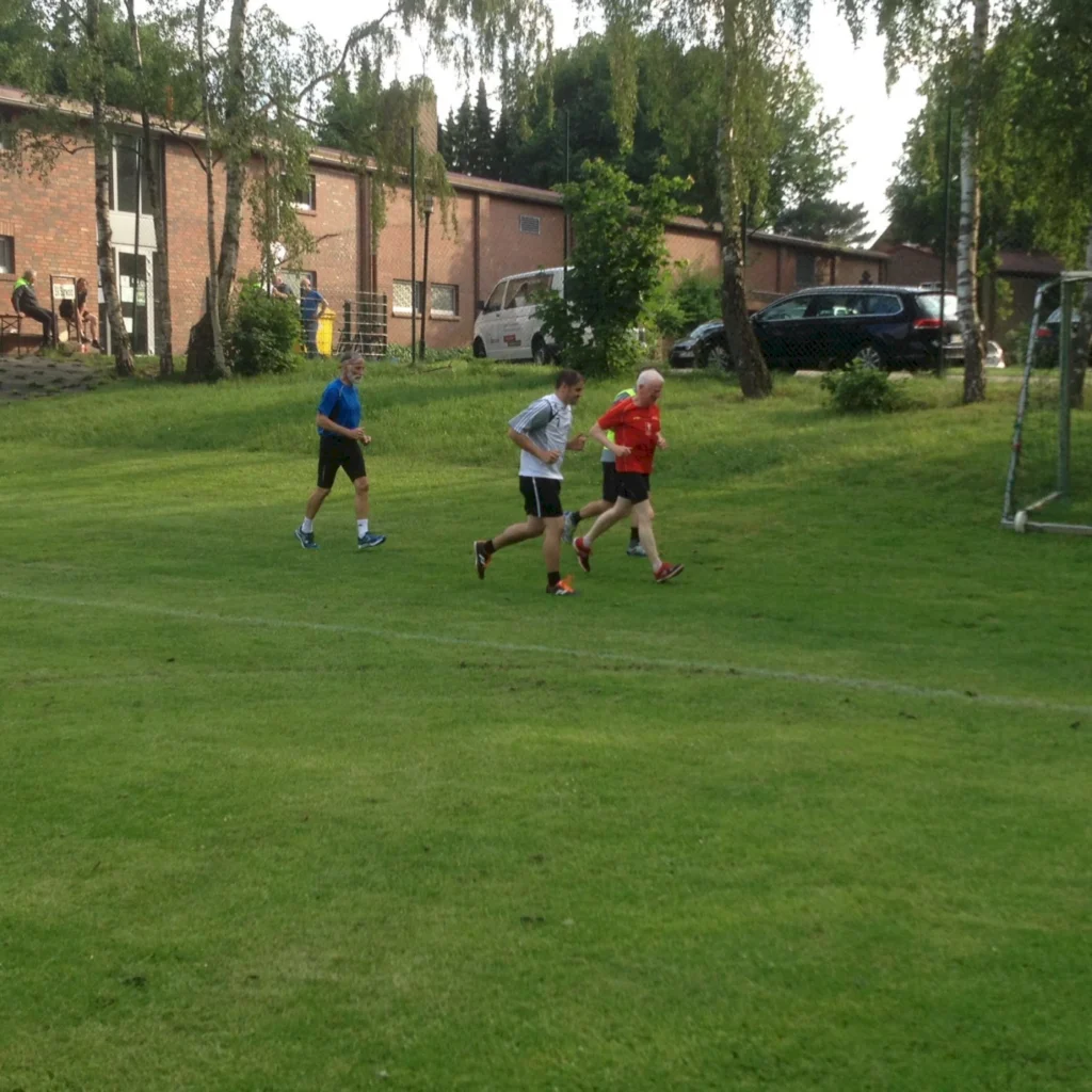 Drei Männer joggen auf einem grünen Sportplatz neben Backsteingebäuden an einem sonnigen Tag.