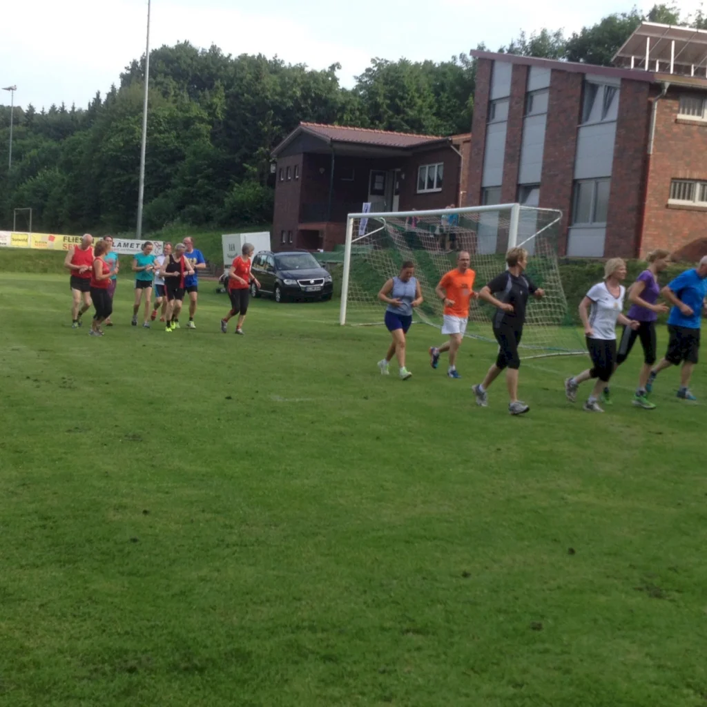 Gruppe joggender Menschen auf einem grünen Sportplatz vor einem Gebäude und Tor, Training im Freien.