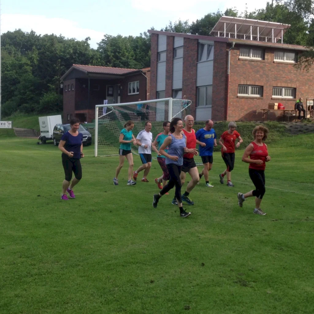 Eine Gruppe von Menschen joggt fröhlich auf einem Sportplatz bei sonnigem Wetter vor einem Vereinsgebäude.