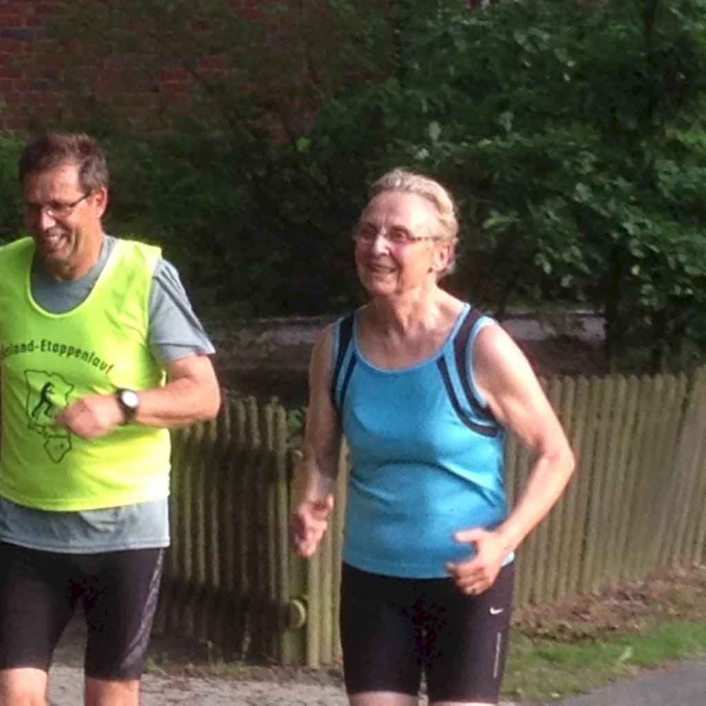 Zwei ältere Menschen in Sportkleidung joggen im Park, lächeln und genießen das sonnige Wetter.