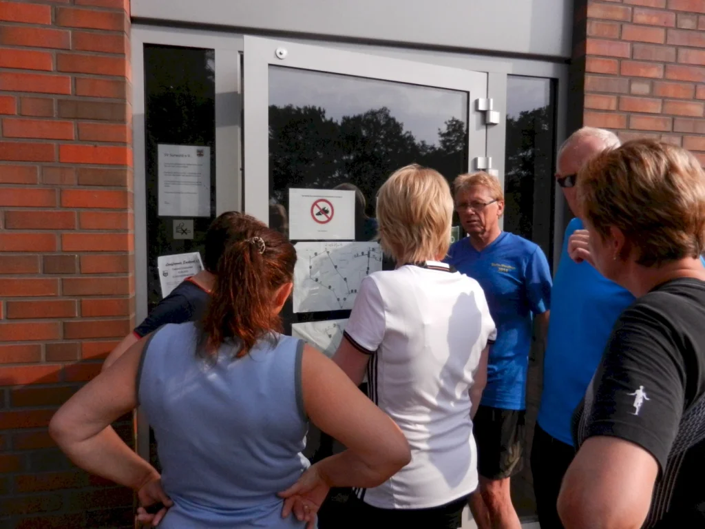 Gruppe von Personen, die an einem Eingang stehen und Informationen auf einem Plakat lesen, während sie sich um einen Aushang versammeln. Die Szene zeigt einen Austausch über die Inhalte des Aushangs, der möglicherweise Regeln oder Hinweise enthält.