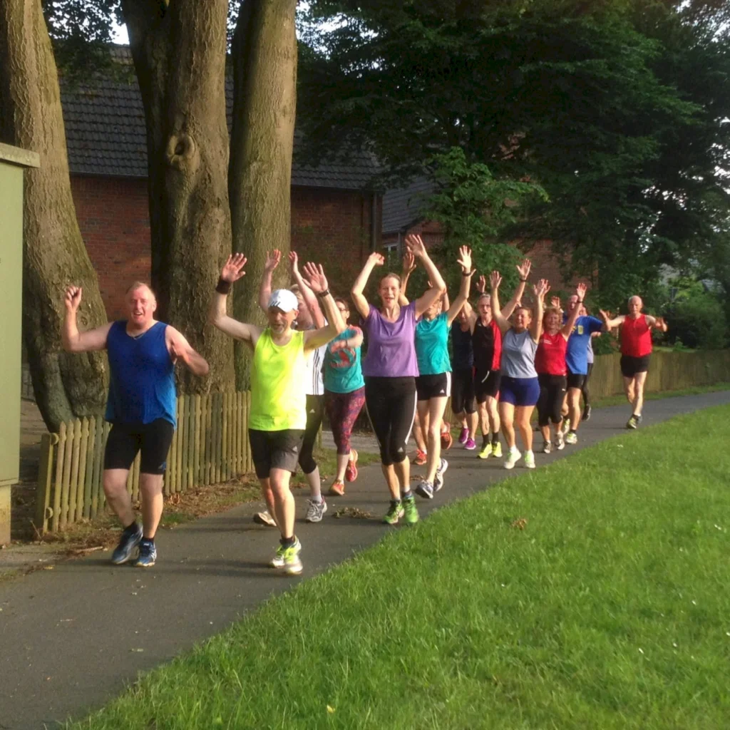 Gruppe von fröhlichen Joggern in bunter Sportkleidung läuft auf einem Weg neben Bäumen und hebt die Arme.