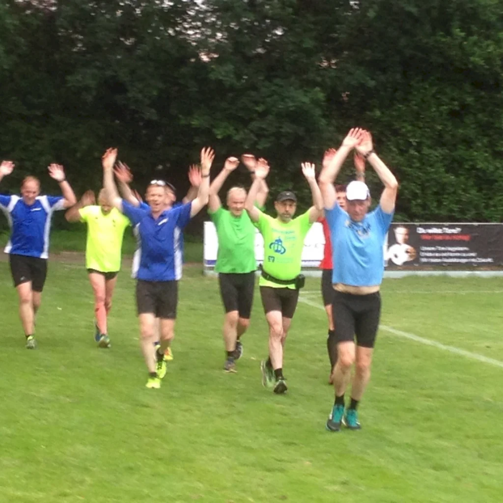 Gruppe von Männern beim Sport auf einem Feld, Arme in die Luft gestreckt, teilweise in blauen und grünen Trikots.