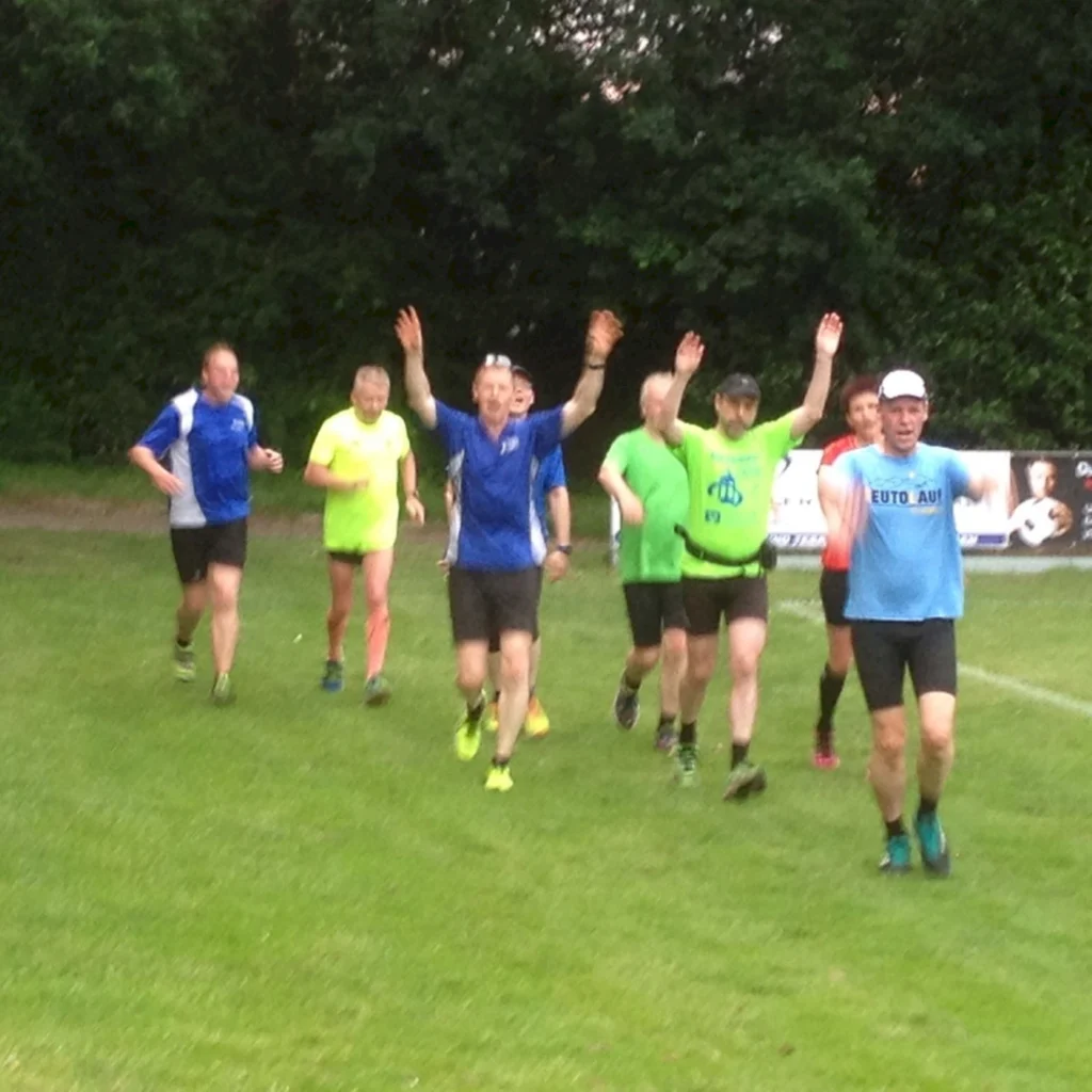 Gruppe von Joggern in bunter Sportkleidung läuft auf grünem Rasen, hebt die Arme in Freude oder Begrüßung.