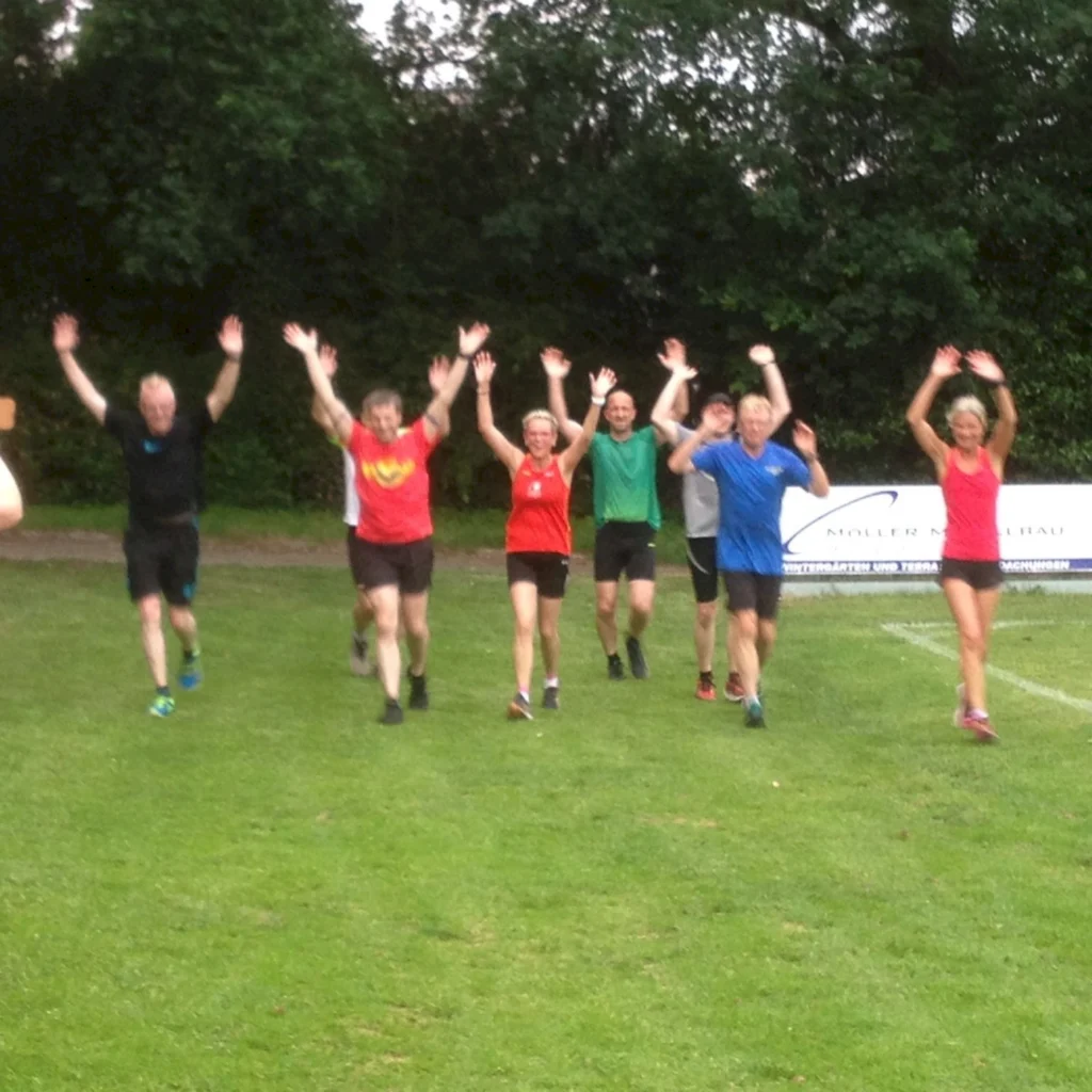 Gruppe von sieben Läufern, die fröhlich mit erhobenen Händen auf einem Rasenfeld laufen, während sie eine Sportveranstaltung oder ein Training absolvieren.