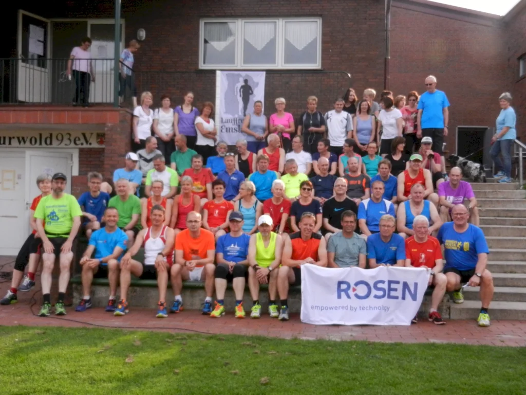 Gruppe von Läufern in bunten Sportkleidung, versammelt vor einem Backsteingebäude, halten ein ROSEN-Banner.