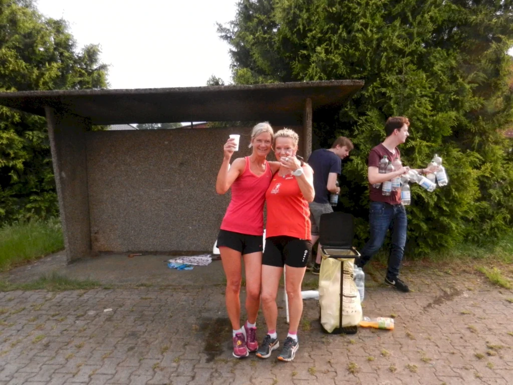 Zwei Frauen in Sportkleidung stehen lächelnd vor einer Bushaltestelle und halten Getränke in der Hand, während im Hintergrund eine Gruppe von Menschen Flaschen mit Wasser vorbereitet.