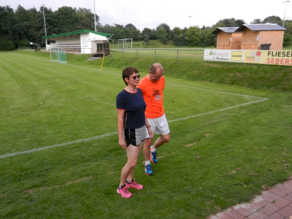 Zwei Personen gehen auf einem Sportplatz spazieren, mit Bänken und Werbung im Hintergrund.