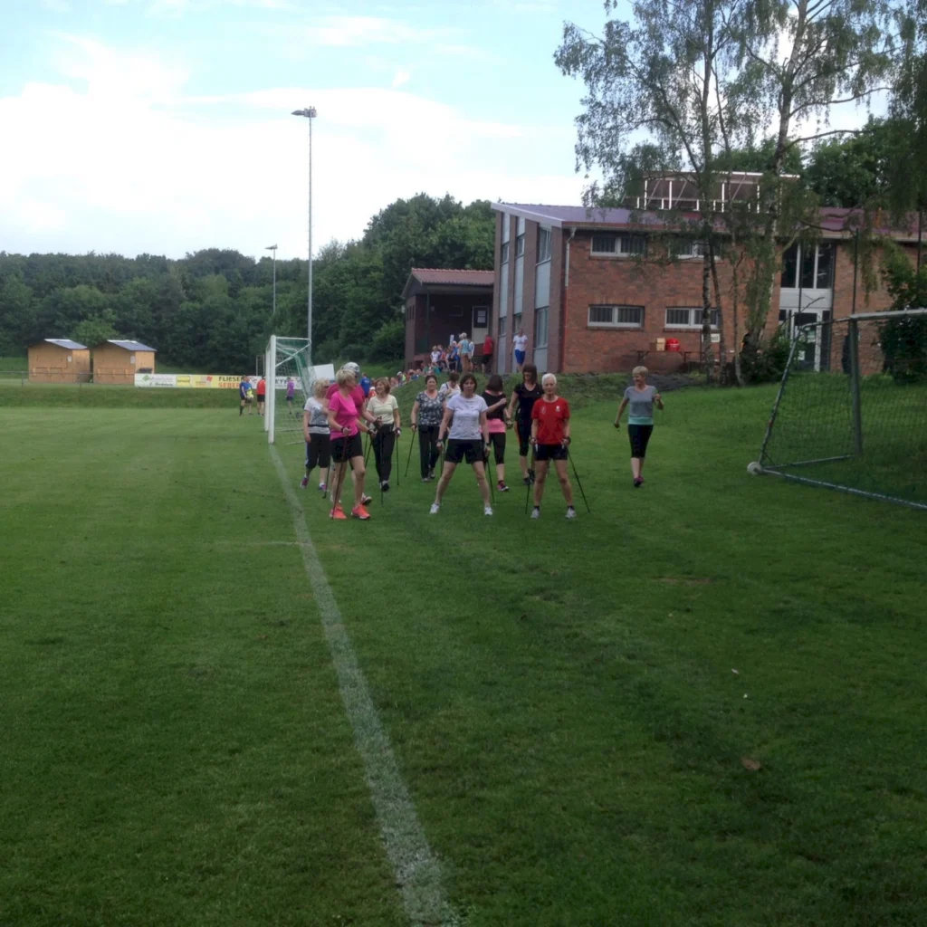 Gruppe von Menschen beim Nordic Walking auf einem Sportplatz vor einem Backsteingebäude im Freien.