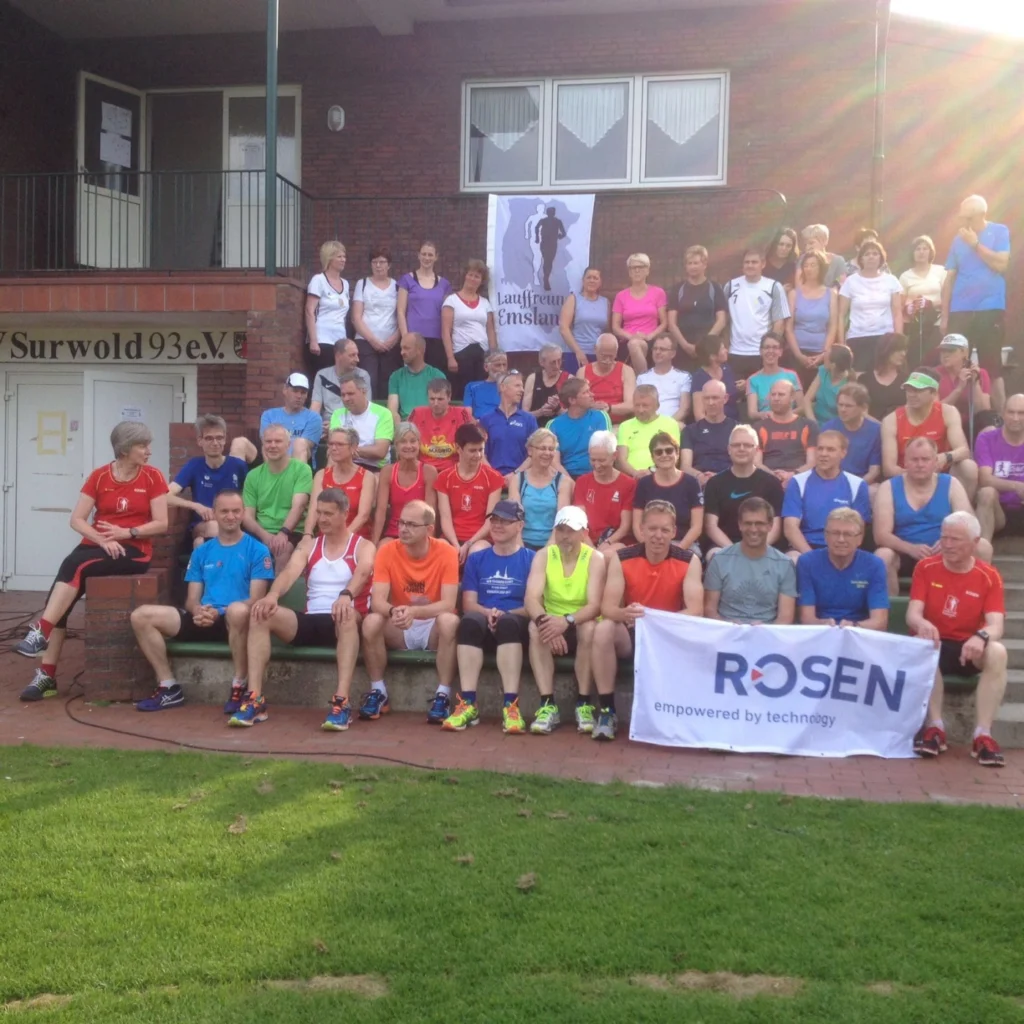 Gruppe von Läufern in bunten Sportoutfits posiert für ein Gruppenfoto vor Vereinsgebäude, Banner von Rosen im Vordergrund.
