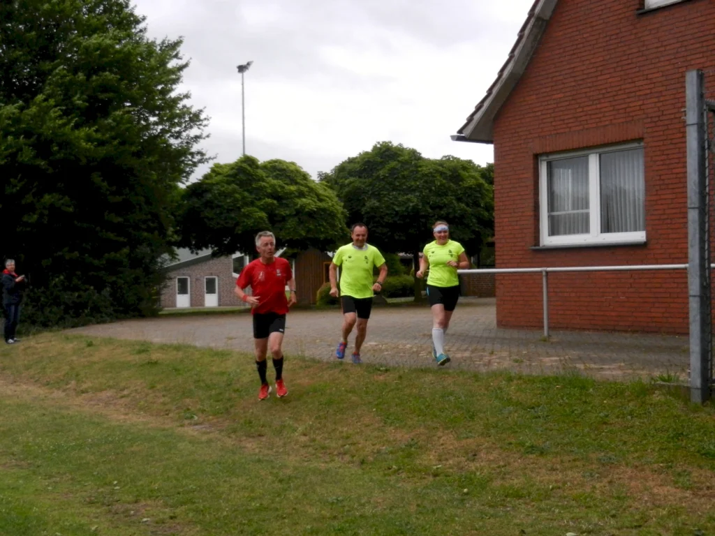 Drei Läufer in leuchtend grünen und roten Shirts joggen auf einem Weg neben einem Gebäude, umgeben von Bäumen und Zuschauern.