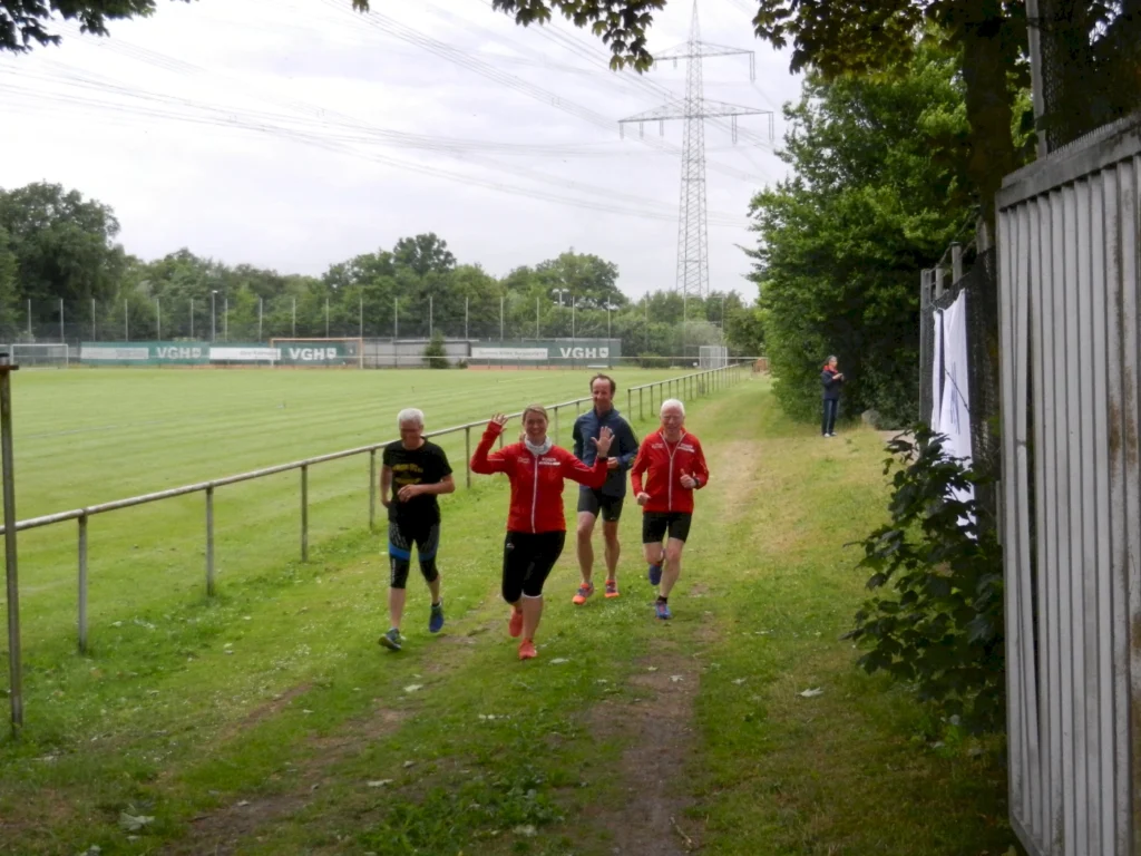 Vier Personen joggen auf einem grasbewachsenen Weg neben einem Fußballfeld unter Stromleitungen.