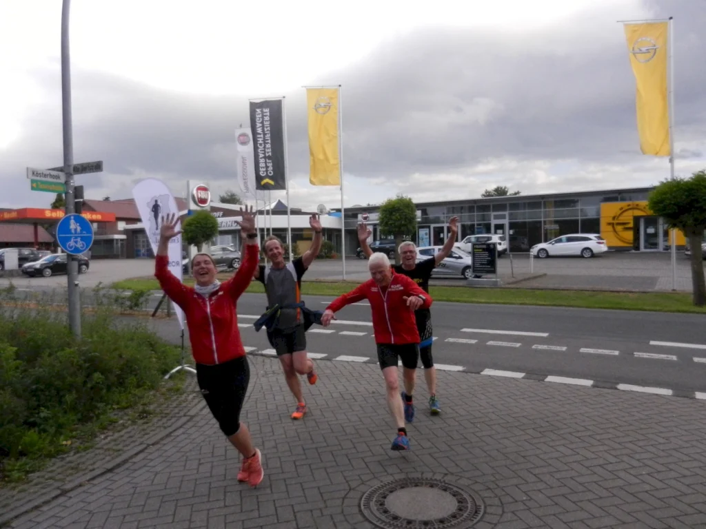 Freudige Läufer überqueren lachend eine Straße bei einer Laufveranstaltung mit Fahnen im Hintergrund.