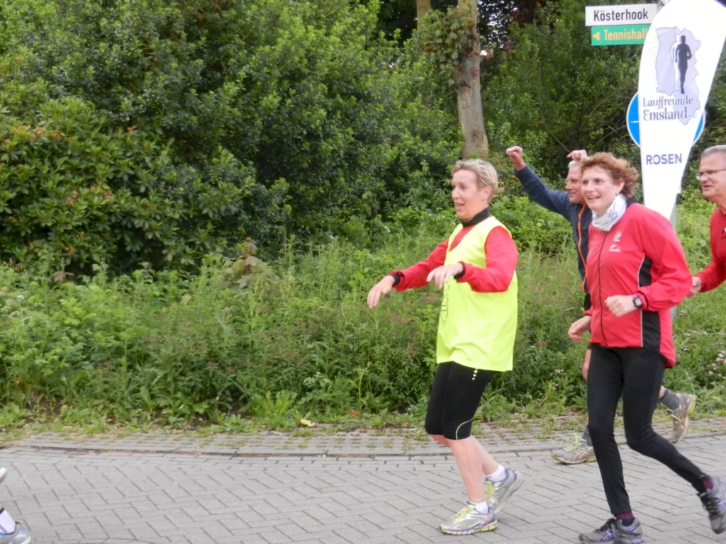 Gruppe von Läufern beim Joggen im Grünen, in sportlicher Kleidung und gut gelaunt, mit einem Wegweiser im Hintergrund.