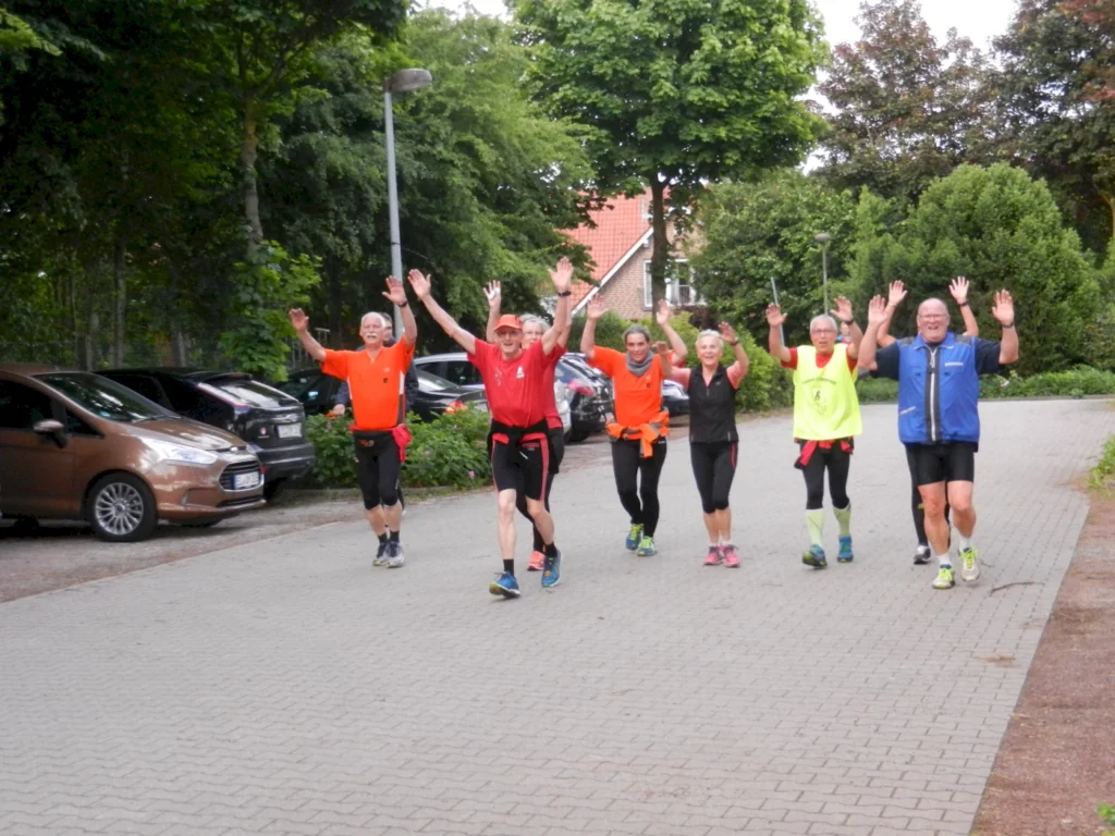 Gruppe älterer Menschen joggt fröhlich mit erhobenen Armen auf einem Bürgersteig vor grüner Baumkulisse.