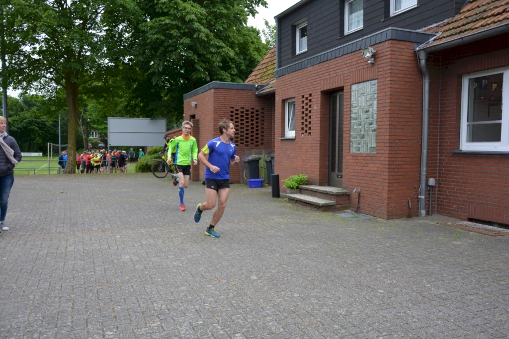 Läufer in Sportkleidung vor Backsteingebäude bei einem Rennen, im Hintergrund Zuschauer auf einem Sportplatz.