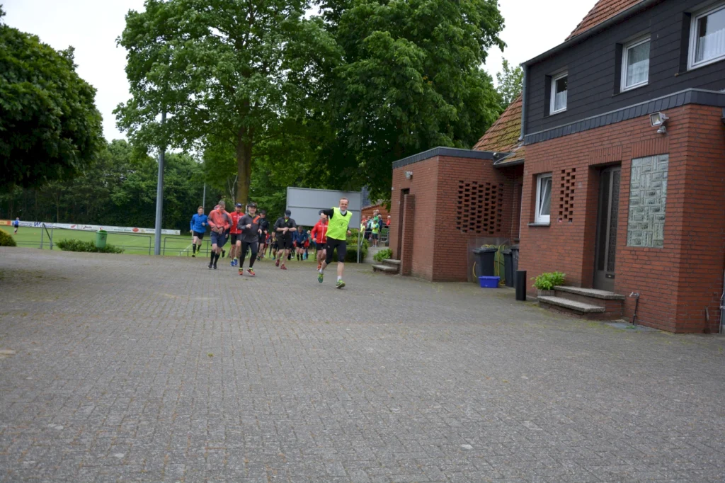 Sportliche Läufergruppe vor Gebäude auf gepflastertem Weg neben Fußballplatz und grünem Park bei bewölktem Himmel.
