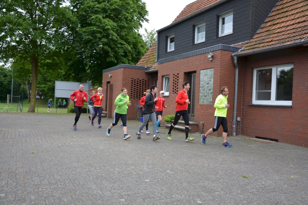 Gruppe von Menschen joggt in Sportkleidung durch eine Wohngegend, umgeben von Bäumen und Backsteingebäuden.