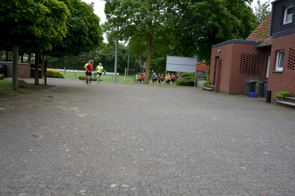 Läufer in bunten Sportoutfits joggen auf einem gepflasterten Weg, umgeben von Bäumen und einem Sportplatz im Hintergrund. Im Hintergrund sind weitere Teilnehmer versammelt.