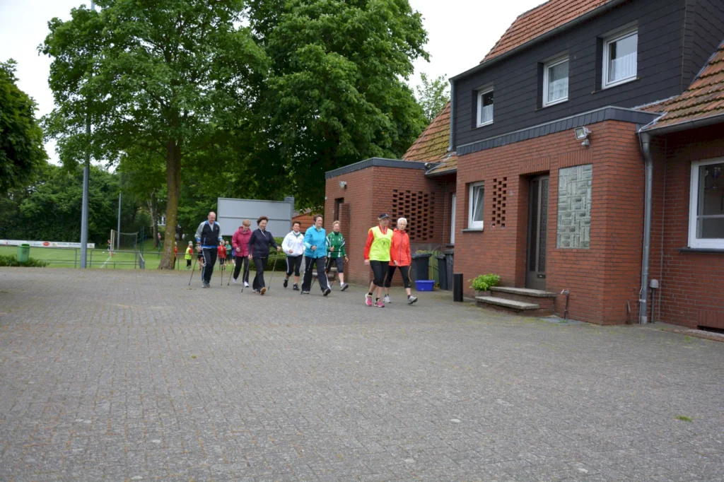 Gruppe von Nordic-Walking-Teilnehmern spaziert vor rotem Backsteinhaus; grüne Bäume im Hintergrund.