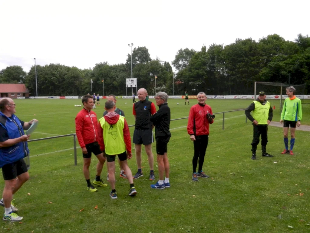 Männer diskutieren auf einem Fußballfeld während eines Trainings, umgeben von Bäumen und Toren.