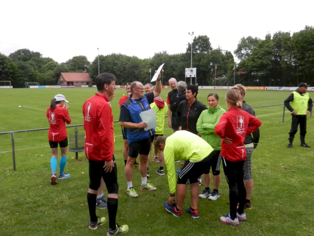 Gruppengespräch von Sportlern auf einem Fußballplatz, mit Teilnehmern in Sportbekleidung, die sich auf eine Trainingseinheit oder einen Wettkampf vorbereiten. Im Hintergrund sind Fußballtore und ein Vereinsgebäude sichtbar.