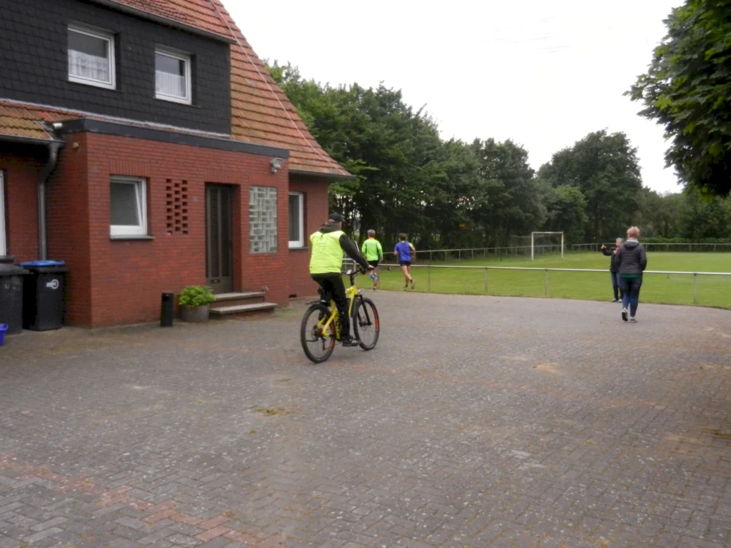 Radfahrer mit gelber Weste vor Backsteingebäude, Läufer auf Weg zum Sportplatz im Hintergrund.