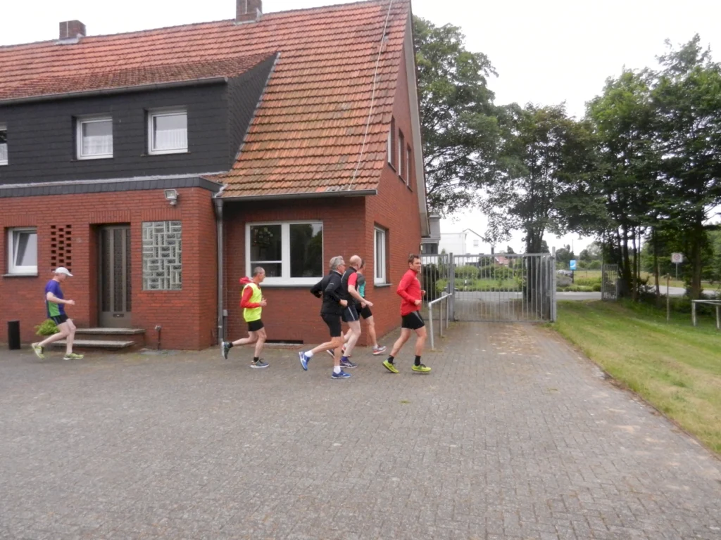 Gruppe von Läufern in bunter Sportkleidung läuft vor einem Backsteinhaus auf einem gepflasterten Weg.